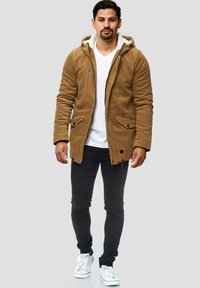 Hombre caminando con una chaqueta de pana marrón con forro de felpa, camiseta blanca de cuello en V, pantalones negros con detalles en las rodillas y zapatillas blancas.