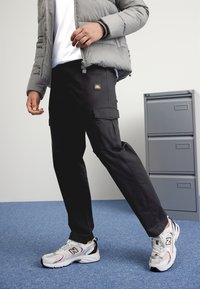 Svarta cargobyxor med flera fickor, tillsammans med en grå vadderad jacka och vita sneakers med röda och svarta detaljer.