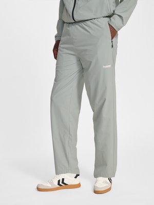 Man draagt lichtgrijze trainingsbroek en bijpassend jack met witte sneakers met zwarte chevronstrepen en rubberen zolen.