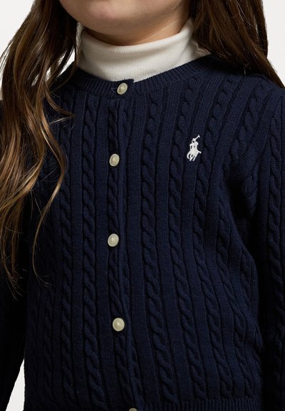 Marinefarbener Strickcardigan mit Rundhals, weißen Knöpfen und gesticktem Logo auf der Brust. Über einem weißen Rollkragenpullover getragen.