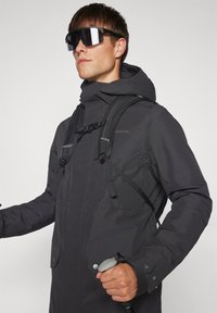 Juodas vandeniui atsparus švarkas su aukštu apykakliu, reguliuojama kapišonu ir priekinėmis kišenėmis. Yra petnešos ir tekstūruota paviršiaus danga.