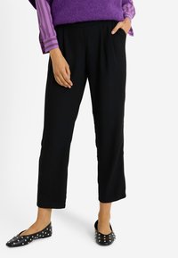 Pantalones negros recortados con un corte relajado y bolsillos laterales, combinados con zapatos planos negros decorados con tachuelas plateadas. El conjunto tiene un aspecto con capas.