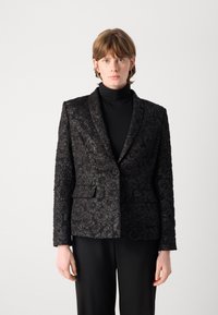 Zwarte blazer met een textuur van een bloemenpatroon, gestructureerde revers, een enkele knoopsluiting en twee voorkruiden. Draagt over een zwarte coltrui.