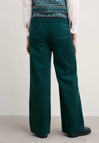Pantalones de pana verdes de pierna ancha con sutiles estrías verticales, dos bolsillos traseros y un dobladillo recto, combinados con una blusa de punto estampada.