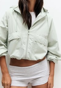 Lichtgroene cropped jas met capuchon, ritssluiting en twee voorzakken. Zachte, lichtgewicht stof met geplooide manchetten. Grijze korte broek eronder.