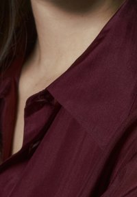 Bordeaux skjorte med en stor krave, der har en glat, let skinnende tekstur og subtil glans. Stoffet virker let og flydende.