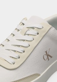 Gros plan d'une basket beige et blanche avec des lacets blancs et le logo "cK" sur un panneau latéral en tissu texturé.
