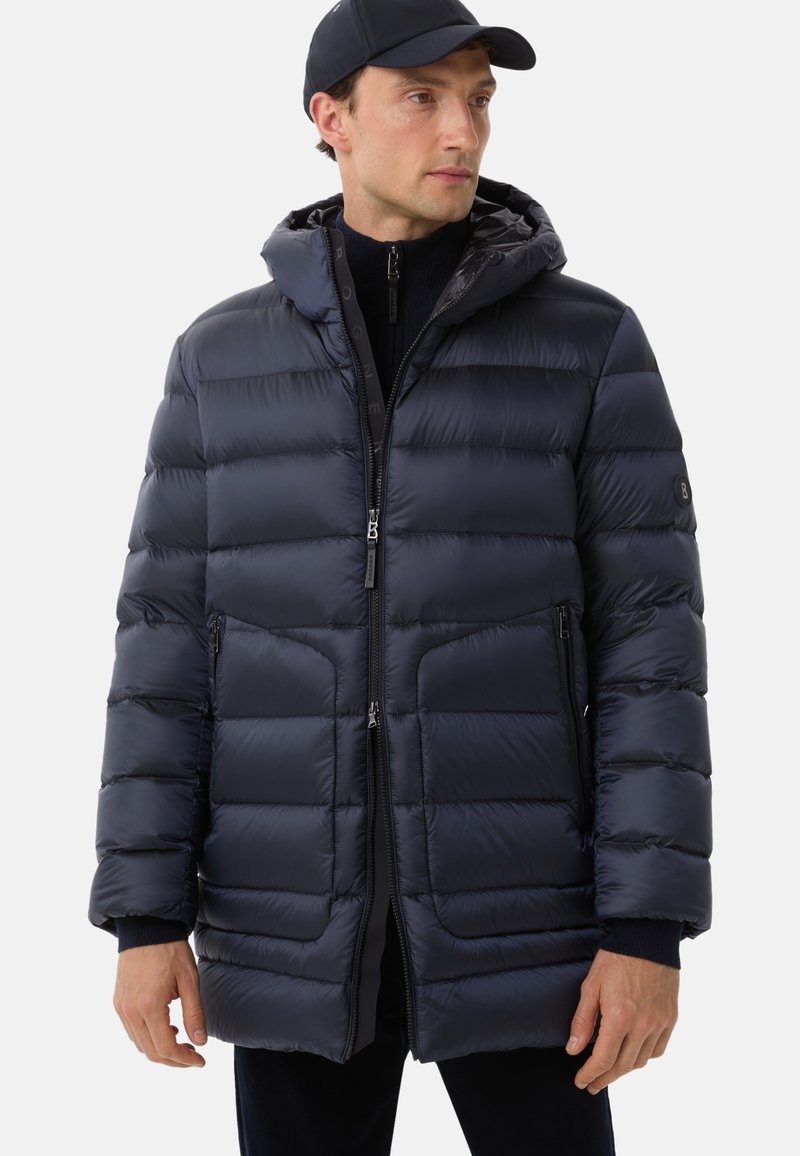 Man wearing navy blue padded hooded winter coat and matching cap, facing slightly right against a plain background.