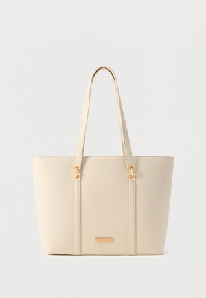 Sac fourre-tout texturé couleur crème, avec longues anses et détails métalliques dorés sur un fond neutre.