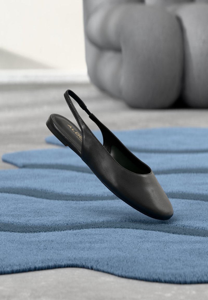 Chaussure plate en cuir noir à bride arrière posée sur un tapis bleu texturé avec un fond gris matelassé.