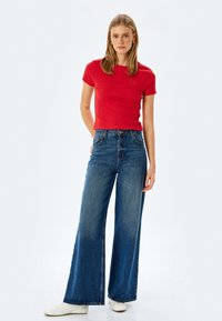 Rød ribbet t-shirt med korte ærmer, parret med højttaljede, vide blå jeans i denim. Modellen har hvide sneakers på.