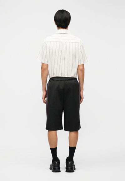 Άνδρας με την πλάτη, φοράει λευκό κοντομάνικο πουκάμισο με λεπτές κάθετες ρίγες, μαύρα σορτς μέχρι το γόνατο με πλαϊνές ρίγες, μαύρες κάλτσες και μαύρα αθλητικά παπούτσια.