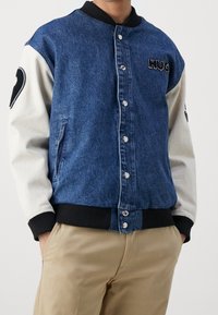 Kurtka dżinsowa z niebieskim korpusem i białymi rękawami, ozdobiona haftem "HUG", z zapięciem na zatrzaski, prążkowanym kołnierzem i bocznymi kieszeniami.