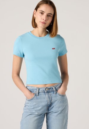 Femme aux cheveux bruns lisses portant un t-shirt court à manches courtes bleu clair et un jean bleu clair taille haute, les mains dans les poches.