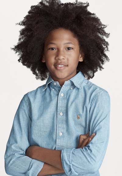 Blå denimskjorte med knappelukking og lange ermer, samt en liten brodert logo på venstre bryst. Skjorten har krage og knappeermer.