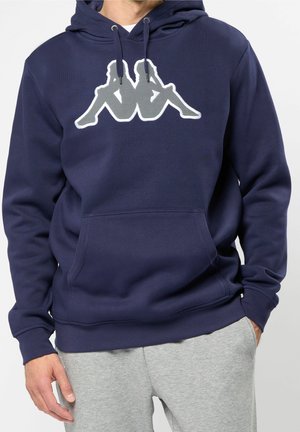 Homme portant un sweat à capuche bleu marine avec un logo de figures blanches et grises assises dos à dos, associé à un pantalon de survêtement gris clair.