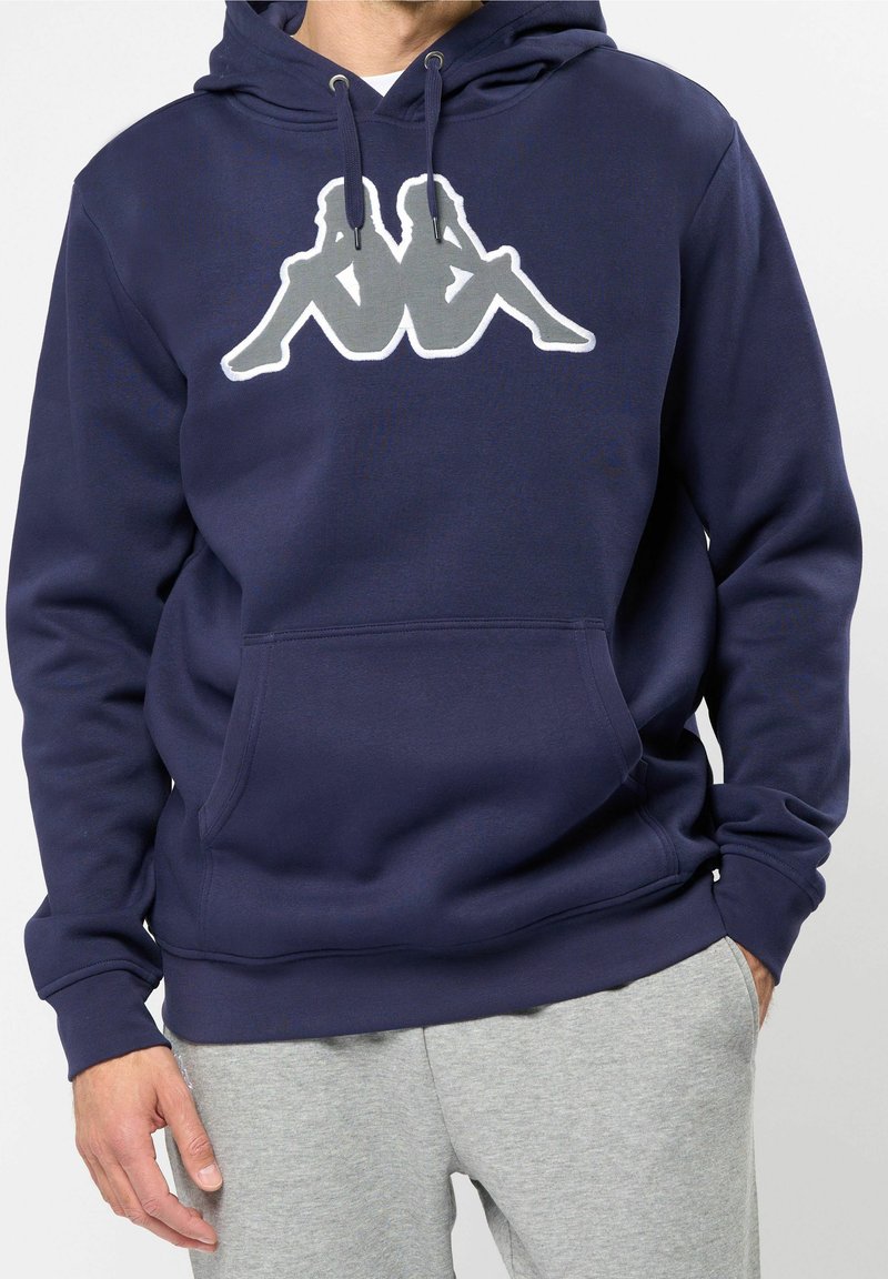 Homme portant un sweat à capuche bleu marine avec un logo de figures blanches et grises assises dos à dos, associé à un pantalon de survêtement gris clair.