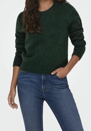 Femme portant un pull en maille vert foncé et un jean bleu en denim, une main dans la poche et l'autre détendue le long du corps.