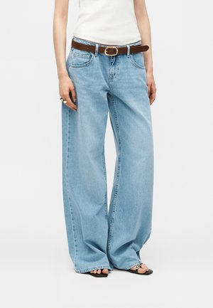 Personne portant un jean bleu clair à jambes larges, une ceinture marron, un haut blanc sans manches et des sandales noires ouvertes à l'avant avec une chaîne décorative en bronze.