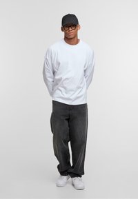 White long-sleeve T-shirt, oversized fit, paired with dark gray loose-fit jeans and white sneakers. Worn with a black cap and glasses.
