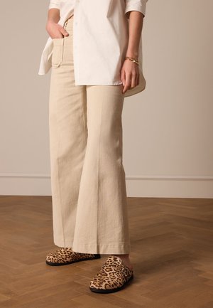 Beige wide-leg pants made of textured fabric, paired with leopard-print clogs featuring a side buckle. White button-up shirt drapes loosely.