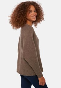 Pull en maille marron avec une coupe décontractée, poignets côtelés et col rond, présenté sur un modèle aux cheveux bouclés, associé à un jean sombre.