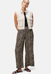 Beige mouwloze utility vest met zakken, gecombineerd met wijde luipaardprintbroeken in gedempte tinten. Zwarte platte sandalen maken de outfit af.