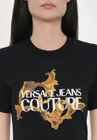 Crna pamučna majica sa višebojnim grafičkim dizajnom i natpisom "VERSACE JEANS COUTURE" te raskošnim zlatnim detaljima.