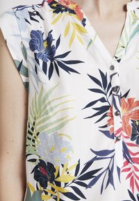 Blusa floral con fondo blanco, que presenta coloridos patrones botánicos. Mangas cortas, frente de botones y detalles de tela texturizada.