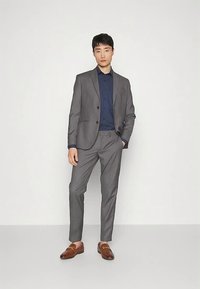 Grå kostym med en figursydd kavaj och byxor, kombinerad med en mörkblå skjorta med prickar. Bruna loafers fullbordar utseendet. Enfärgad bakgrund.