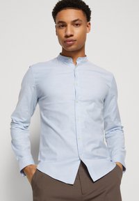 Camicia a maniche lunghe azzurro chiaro con colletto alla coreana, chiusura a bottoni e tessuto leggermente testurizzato. La camicia ha una vestibilità slim e un orlo arrotondato.