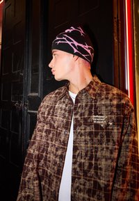 Brown plaid shirt with a black beanie featuring pink abstract patterns. The shirt has a subtle logo on the left chest area.