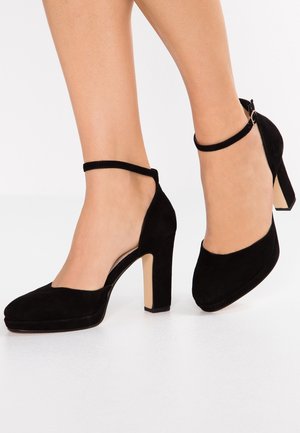 Nogi w czarnych zamszowych butach na wysokim obcasie z paskami wokół kostki i grubymi obcasami na białej powierzchni.
