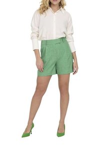 Pantalones cortos verdes con textura, de tiro alto y pliegues frontales, combinados con una camisa blanca de botones y zapatos de tacón verdes.