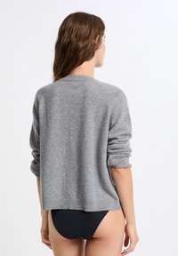 Femme aux longs cheveux bruns portant un pull gris et des sous-vêtements noirs, se tenant de dos contre un fond blanc.