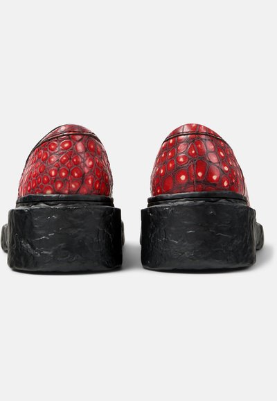 Coppia di scarpe rosse in pelle testurizzata con suole nere spesse e irregolari, viste da dietro su uno sfondo semplice.