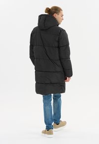 Sort pufferjakke med hætte, der har quiltede sektioner, knapmanchetter og en lang design. Båret over blå jeans og beige sneakers.