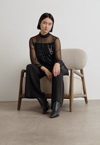 Schwarzes glitzerndes Oberteil mit durchsichtigen Ärmeln, kombiniert mit schwarzen weit geschnittenen Hosen und spitzen Ankle-Boots, sitzt auf einem hellen gepolsterten Stuhl.