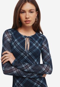 Meriino ruudullinen pitkähihainen mekko, jossa on pyöreä kaula-aukko ja etuosassa solmitys. Ominaisuuksina vaaleansinisiä, valkoisia ja ruskeita yksityiskohtia. Pehmeä, tiukasti istuva kangas.