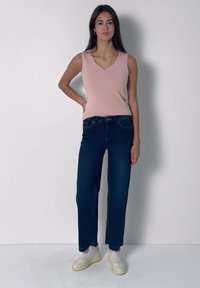 Femme debout contre un mur blanc portant un haut rose sans manches, un jean bleu foncé, des baskets blanches, une main sur la hanche.