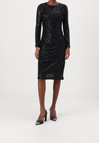 Robe noire en sequins avec des manches longues, longueur genoux, col rond et coupe ajustée. Assortie à des chaussures à talons hauts noires avec des accents en argent.