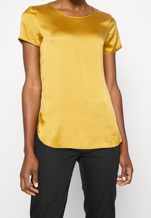 Kvinde iført en kortærmet, skinnende sennepsgul bluse kombineret med sorte bukser, stående mod en ensfarvet hvid baggrund.