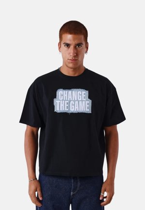 Joven con cabello corto y castaño que lleva una camiseta negra con la frase "CHANGE THE GAME" en letras azul claro, combinada con jeans oscuros.