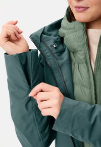 Due giacche sovrapposte: l'esterno è di un verde scuro uniforme, tessuto liscio; l'interno è di un verde oliva chiaro, trapuntato, con un colletto alto e dettagli con zip.
