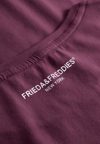 Bordeaux katoenen stof met een gladde textuur; heeft witte bedrukte tekst "FRIEDA & FREDDIES NEW YORK" nabij de halslijn.
