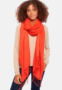 Écharpe rouge en tissu doux, présentant des bords frangés et un design drapé, portée sur un pull beige clair.