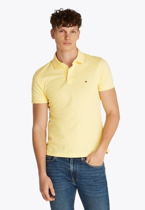 Ung mand iført en lysegul kortærmet poloshirt og blå jeans, stående med den ene hånd i lommen mod en ensfarvet baggrund.