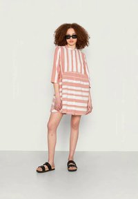 Vestido de manga curta em listras corais e brancas, com um ajuste solto e cintura franzida. Combinado com sandálias pretas e óculos de sol.