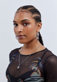 Top em malha preta com estampagem gráfica colorida. O modelo usa brincos de argola prateados e um colar de corrente prateado com um pendente. Textura de pele suave.