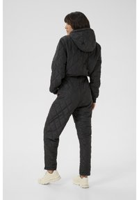 Schwarzer gesteppter Overall mit Kapuze, der eine taillierte Taille, lange Ärmel und elastische Bündchen aufweist. Getragen mit hellen Sneakern.